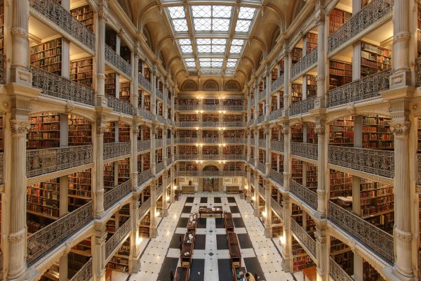 Interior of the George Peabody Library in Baltimore. A wide-angle view of multiple levels of a library. Black and white checkered floors, with various balconies on the sides filled with rows of books. An open skylight illuminates the scene alongside lights on each level