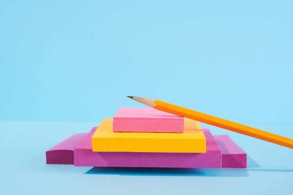 A pencil sits a top purple, yellow, and pink sticky notes in front of a light blue background